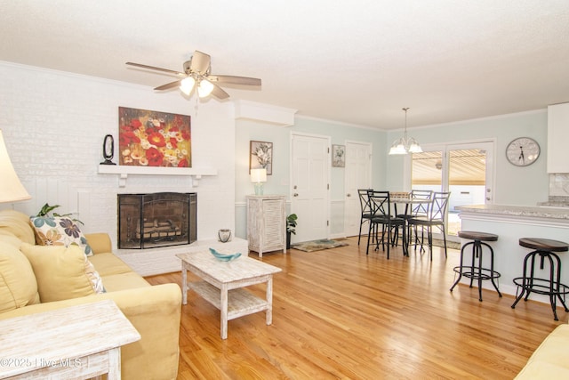 living room with light wood-style floors, a brick fireplace, crown molding, and ceiling fan with notable chandelier