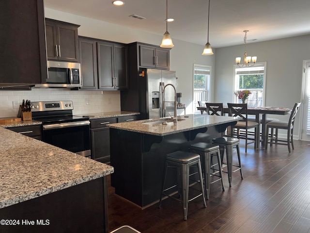 kitchen featuring appliances with stainless steel finishes, decorative light fixtures, dark wood-type flooring, light stone counters, and a kitchen island with sink