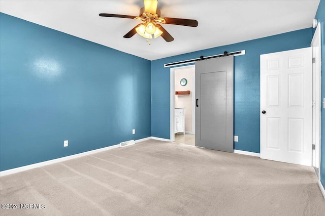 unfurnished bedroom featuring a barn door, light colored carpet, ceiling fan, and ensuite bathroom