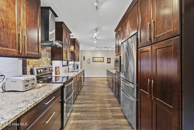 kitchen featuring wall chimney range hood, light stone counters, appliances with stainless steel finishes, light hardwood / wood-style flooring, and sink