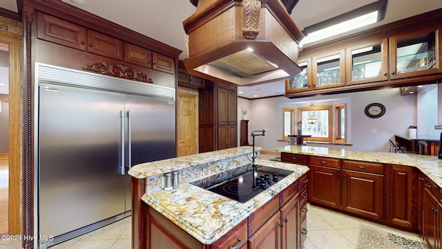 kitchen with custom exhaust hood, an island with sink, built in fridge, black electric stovetop, and light stone countertops