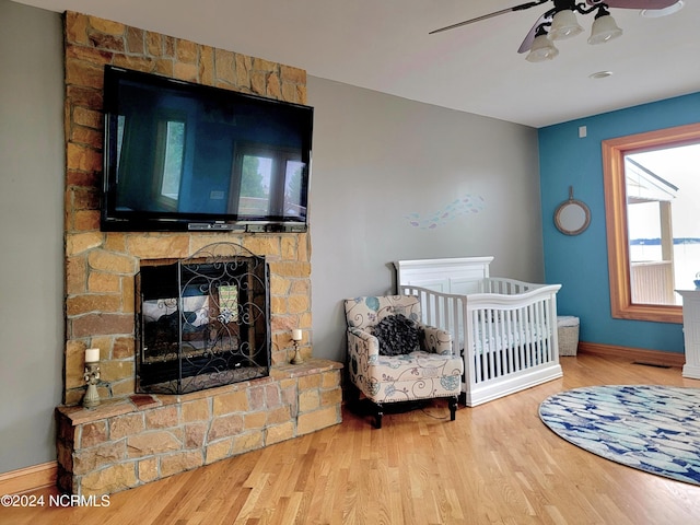 bedroom with a nursery area, ceiling fan, wood-type flooring, and a stone fireplace