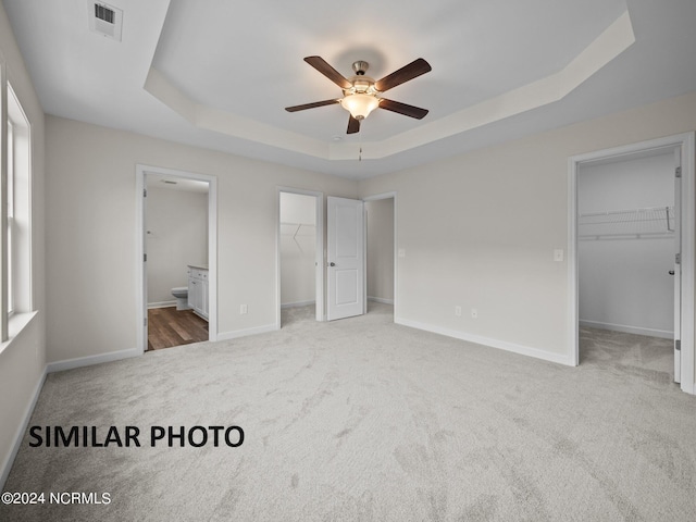 unfurnished bedroom featuring ceiling fan, a tray ceiling, carpet floors, a walk in closet, and ensuite bath