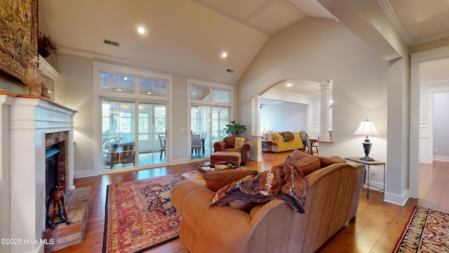 living room featuring crown molding, decorative columns, high vaulted ceiling, light hardwood / wood-style floors, and a brick fireplace