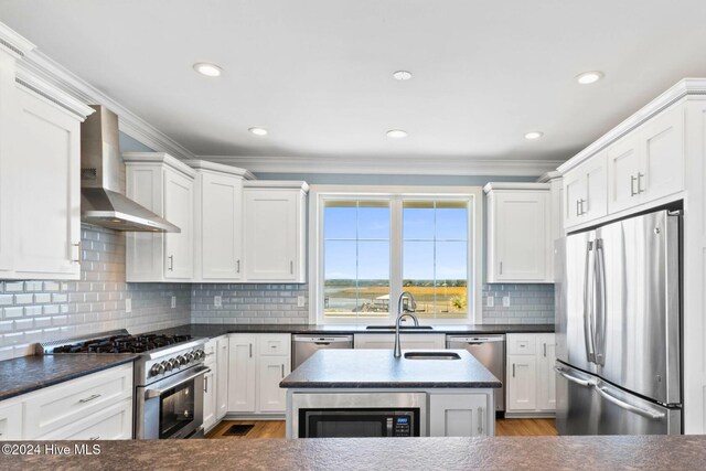 kitchen featuring stainless steel appliances, wall chimney exhaust hood, ornamental molding, a kitchen island with sink, and light hardwood / wood-style flooring