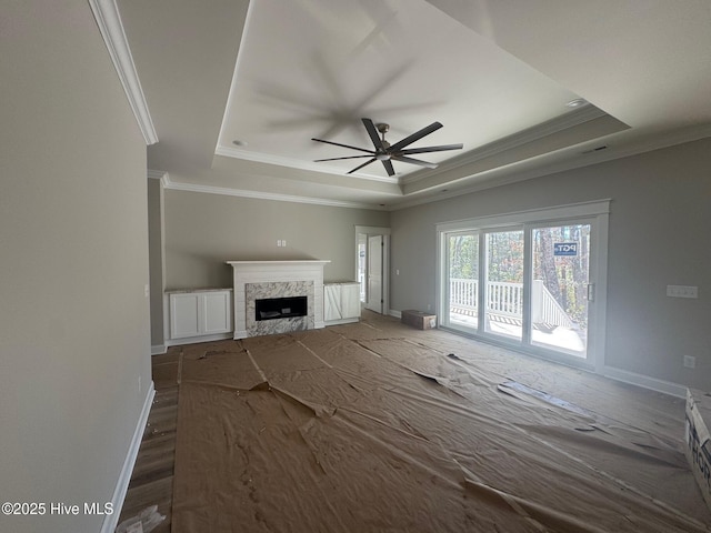 unfurnished living room with a raised ceiling, ornamental molding, a high end fireplace, a ceiling fan, and baseboards