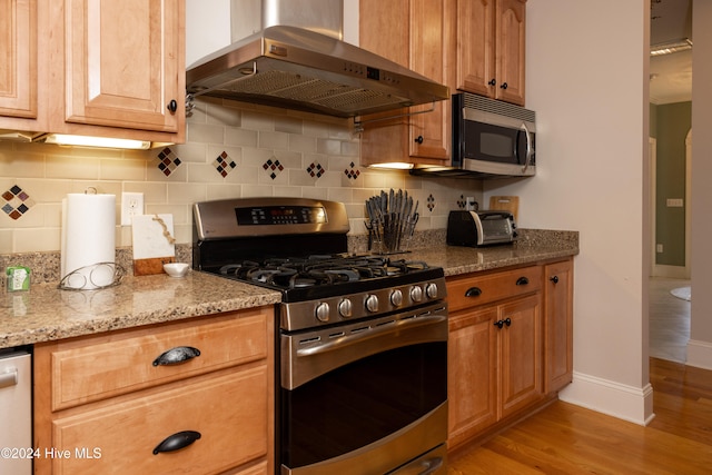 kitchen with wall chimney range hood, light hardwood / wood-style flooring, light stone countertops, appliances with stainless steel finishes, and tasteful backsplash