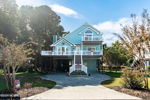 raised beach house with concrete driveway, a front yard, a chimney, a balcony, and a carport