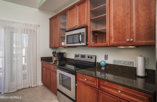 kitchen featuring stainless steel appliances, dark stone countertops, glass insert cabinets, light tile patterned floors, and brown cabinets