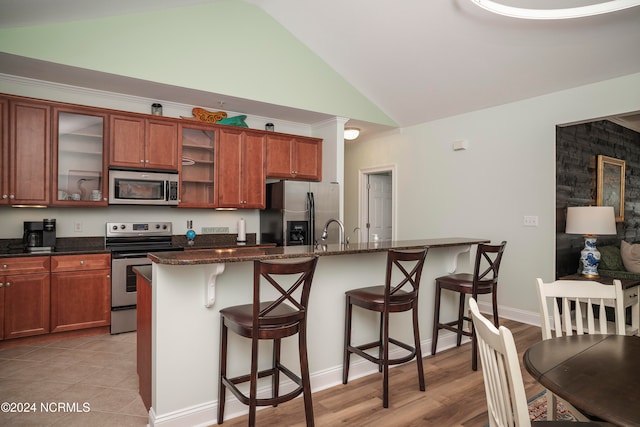 kitchen with a breakfast bar area, appliances with stainless steel finishes, dark stone counters, vaulted ceiling, and light hardwood / wood-style flooring