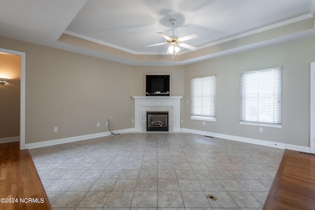 unfurnished living room featuring a tiled fireplace, a ceiling fan, a tray ceiling, and crown molding