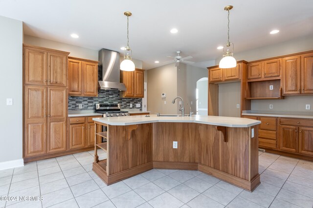 kitchen with sink, stainless steel range oven, ceiling fan, wall chimney exhaust hood, and a kitchen island with sink