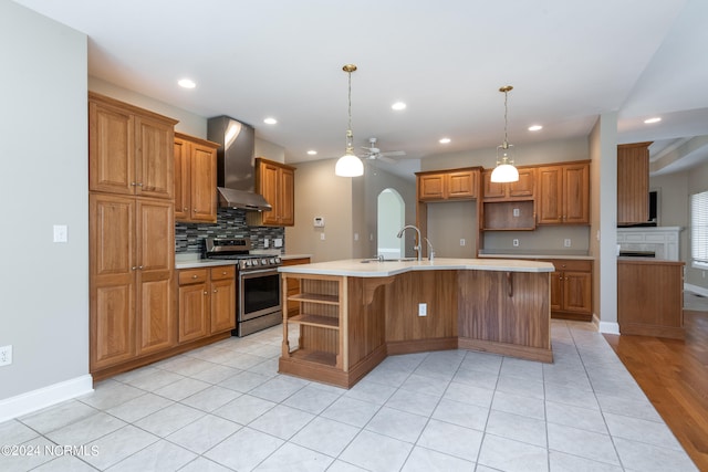 kitchen with wall chimney exhaust hood, an island with sink, pendant lighting, stainless steel range oven, and ceiling fan