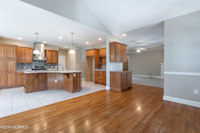 kitchen with a kitchen island with sink, pendant lighting, stainless steel range, wall chimney exhaust hood, and light hardwood / wood-style floors