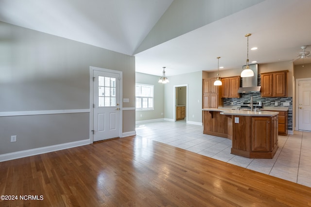 kitchen with a kitchen bar, light wood-type flooring, wall chimney exhaust hood, decorative light fixtures, and a center island with sink