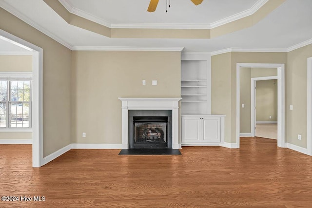 unfurnished living room featuring a raised ceiling, built in shelves, wood-type flooring, and ornamental molding