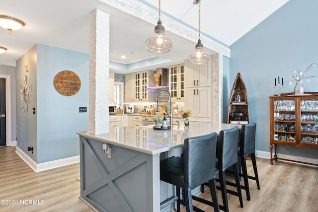 kitchen with wall chimney range hood, light stone countertops, a breakfast bar area, hanging light fixtures, and light hardwood / wood-style flooring