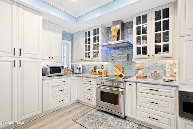 kitchen with wall chimney range hood, stainless steel stove, white cabinets, light hardwood / wood-style floors, and tasteful backsplash