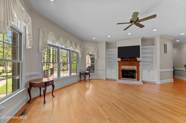 living area with crown molding, a glass covered fireplace, ceiling fan, baseboards, and visible vents