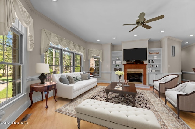 living area featuring a ceiling fan, a glass covered fireplace, light wood finished floors, ornamental molding, and visible vents