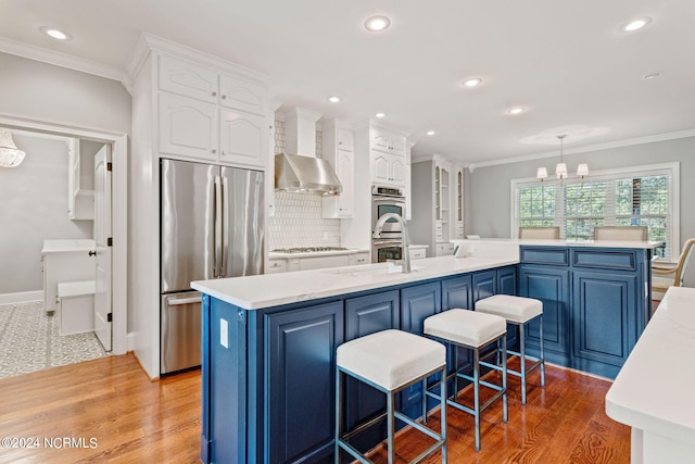 kitchen with wall chimney range hood, hanging light fixtures, stainless steel appliances, an island with sink, and blue cabinets