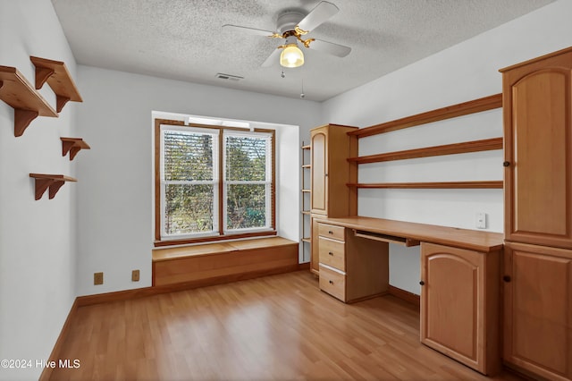 unfurnished office featuring a textured ceiling, light wood-style flooring, visible vents, a ceiling fan, and built in study area