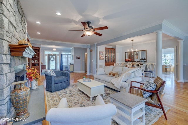 living room featuring light wood-type flooring, ceiling fan with notable chandelier, a stone fireplace, ornate columns, and ornamental molding