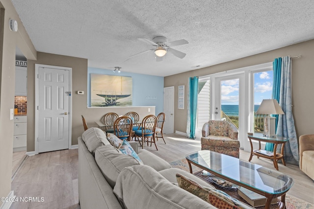 living area with ceiling fan, light wood-style flooring, a textured ceiling, a water view, and baseboards