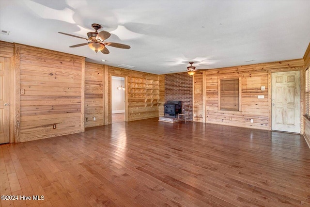 unfurnished living room with ceiling fan, a wood stove, dark wood-type flooring, and wooden walls