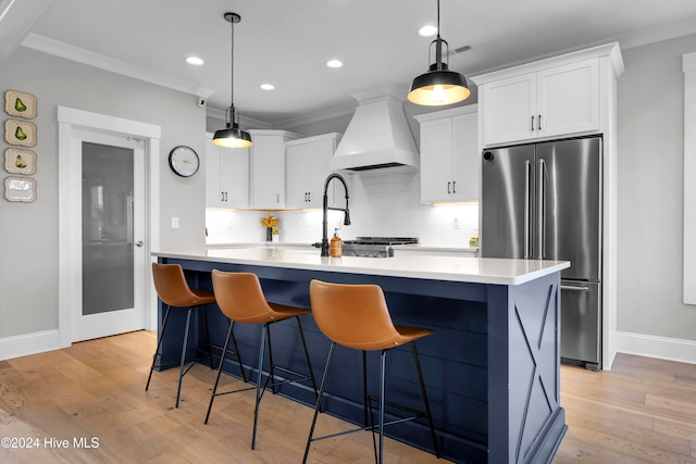 kitchen featuring custom exhaust hood, an island with sink, high quality fridge, ornamental molding, and white cabinets