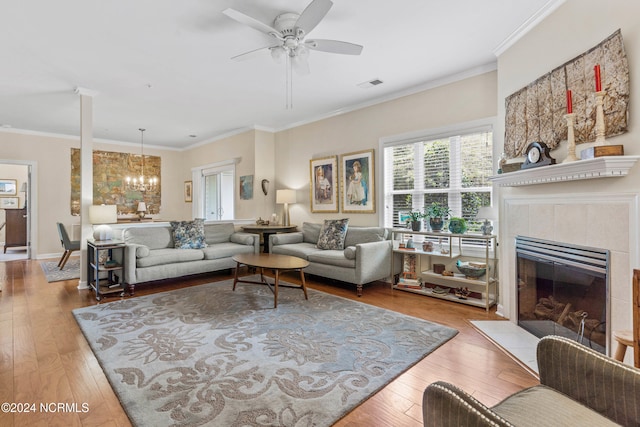 living room featuring wood-type flooring, a tiled fireplace, ceiling fan with notable chandelier, and ornamental molding