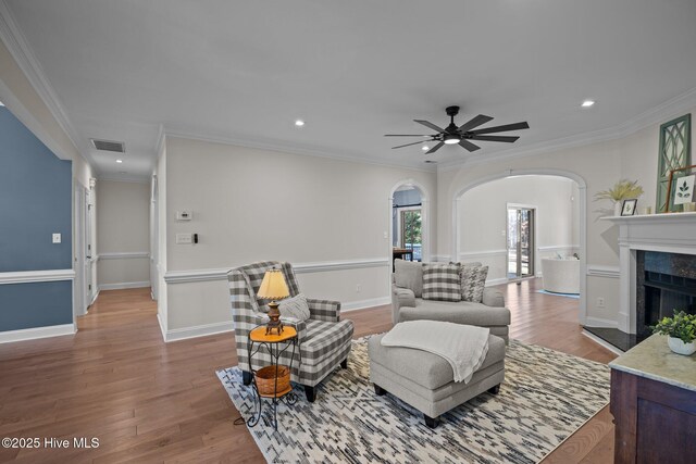 living area with arched walkways, light wood-style flooring, ceiling fan, crown molding, and a high end fireplace