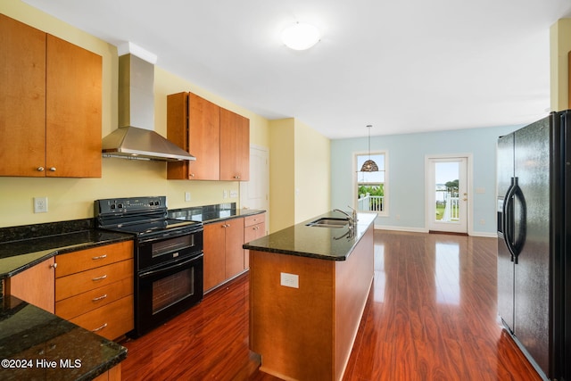 kitchen with dark wood-type flooring, wall chimney range hood, an island with sink, decorative light fixtures, and black appliances