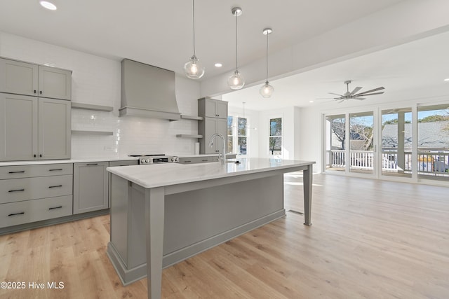 kitchen with an island with sink, premium range hood, ceiling fan, light hardwood / wood-style flooring, and backsplash