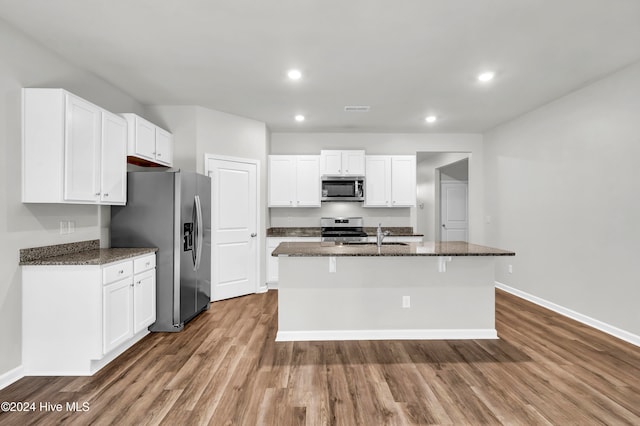 kitchen featuring hardwood / wood-style flooring, stainless steel appliances, dark stone counters, a center island with sink, and white cabinets