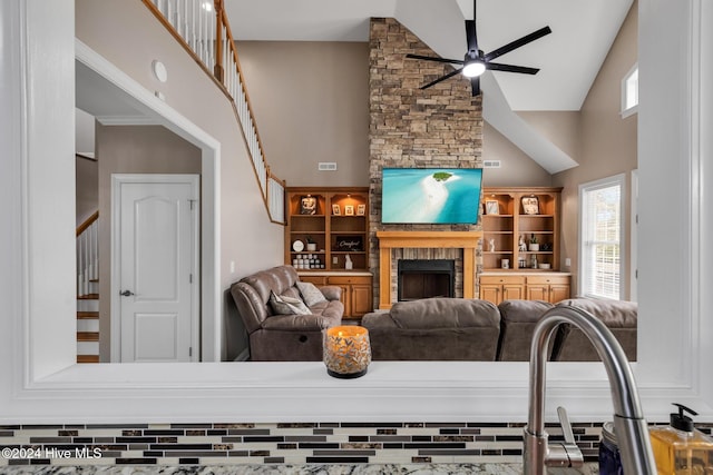 living room with high vaulted ceiling, built in features, a stone fireplace, and ceiling fan