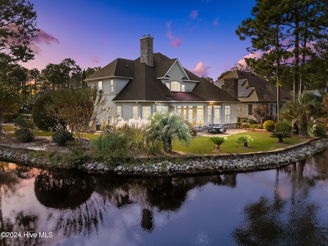 back house at dusk with a water view, a patio area, and a lawn