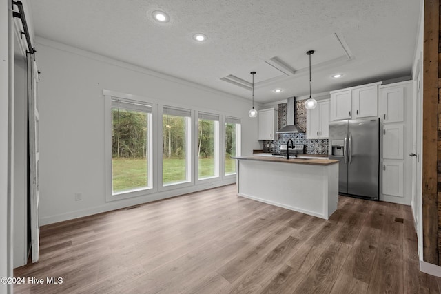 kitchen featuring white cabinetry, hanging light fixtures, wall chimney range hood, stainless steel refrigerator with ice dispenser, and a center island with sink