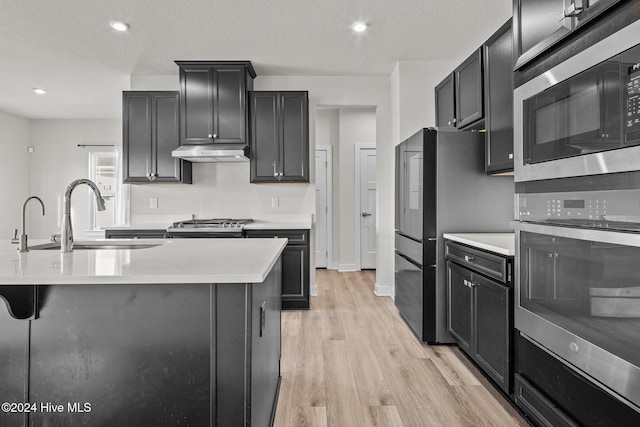 kitchen featuring appliances with stainless steel finishes, a textured ceiling, sink, a center island with sink, and light hardwood / wood-style flooring