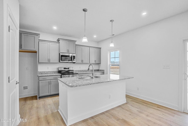 kitchen with sink, hanging light fixtures, appliances with stainless steel finishes, light stone countertops, and a kitchen island with sink