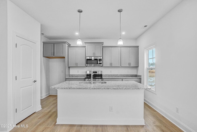 kitchen with gray cabinetry, a center island with sink, appliances with stainless steel finishes, pendant lighting, and light stone countertops