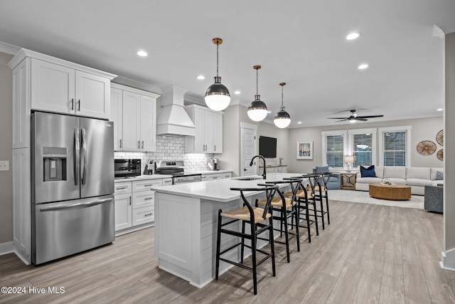 kitchen with hanging light fixtures, a center island with sink, white cabinets, custom range hood, and appliances with stainless steel finishes