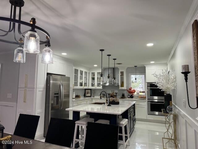 kitchen featuring appliances with stainless steel finishes, a breakfast bar, wall chimney range hood, a center island with sink, and white cabinetry