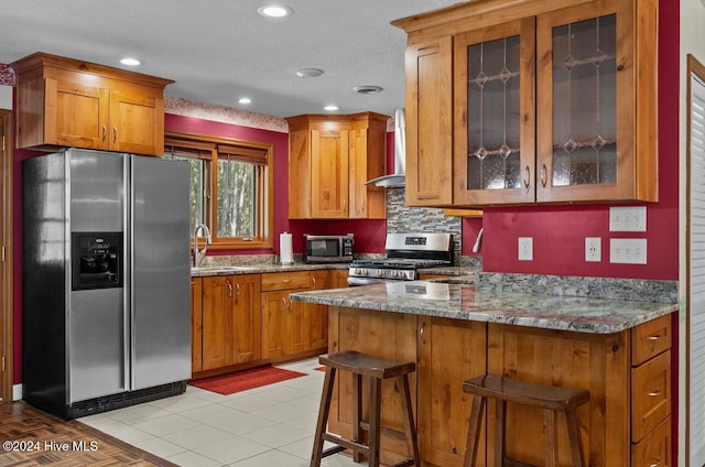 kitchen featuring wall chimney exhaust hood, stainless steel appliances, kitchen peninsula, stone countertops, and a breakfast bar area