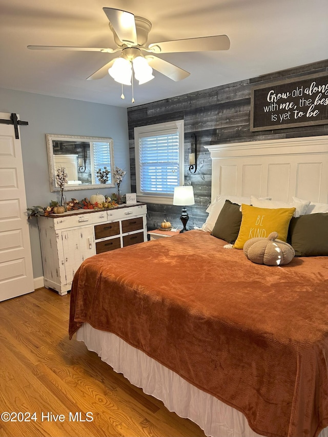 bedroom with ceiling fan, a barn door, wooden walls, and light hardwood / wood-style flooring