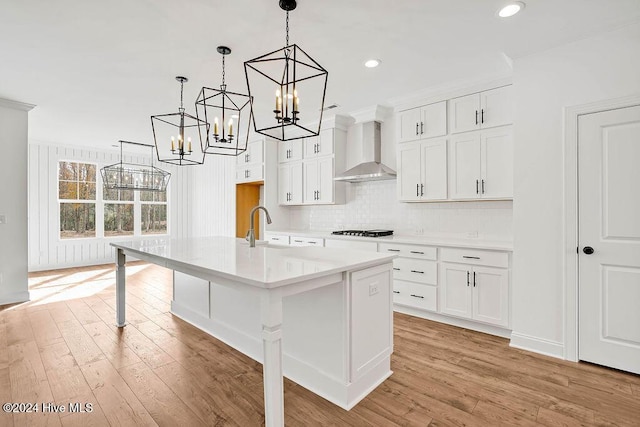 kitchen featuring a center island with sink, white cabinets, pendant lighting, and wall chimney range hood