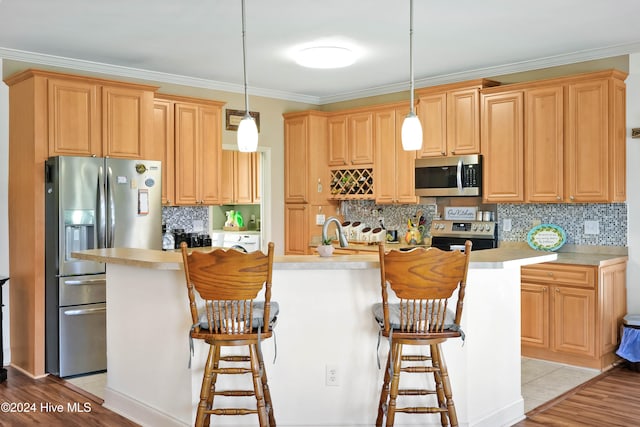 kitchen featuring crown molding, decorative light fixtures, a kitchen island with sink, appliances with stainless steel finishes, and light wood-type flooring