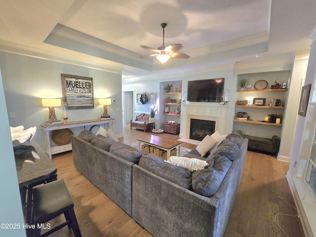 living area with built in shelves, a fireplace with flush hearth, wood finished floors, crown molding, and a raised ceiling