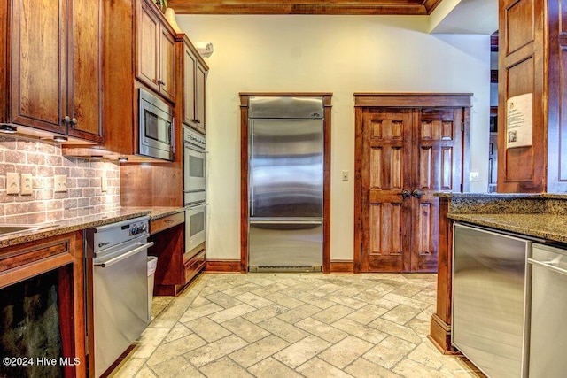 kitchen with built in appliances, brick floor, baseboards, backsplash, and dark stone counters