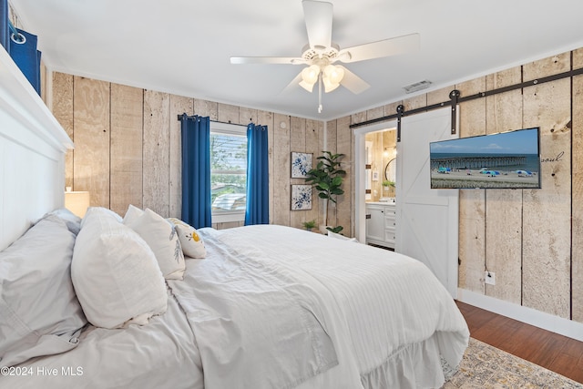 bedroom with connected bathroom, a barn door, wooden walls, hardwood / wood-style floors, and ceiling fan